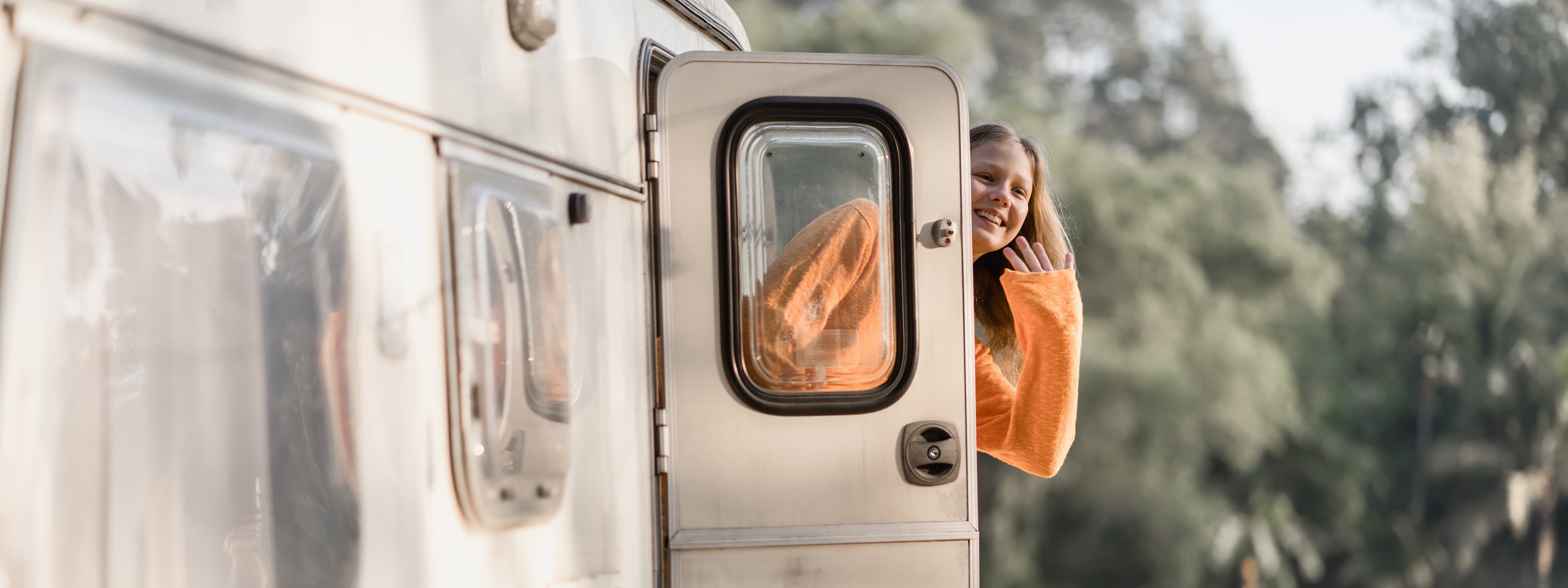 Junges Mädchen winkt fröhlich aus Wohnwagenfenster - Symbol für Familienglück beim Camping