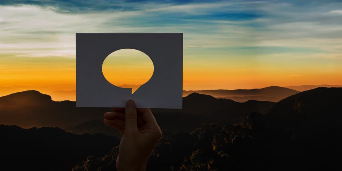 Hand hält Sprechblasen-Silhouette vor Berglandschaft bei Sonnenuntergang als Symbol für Corporate Language