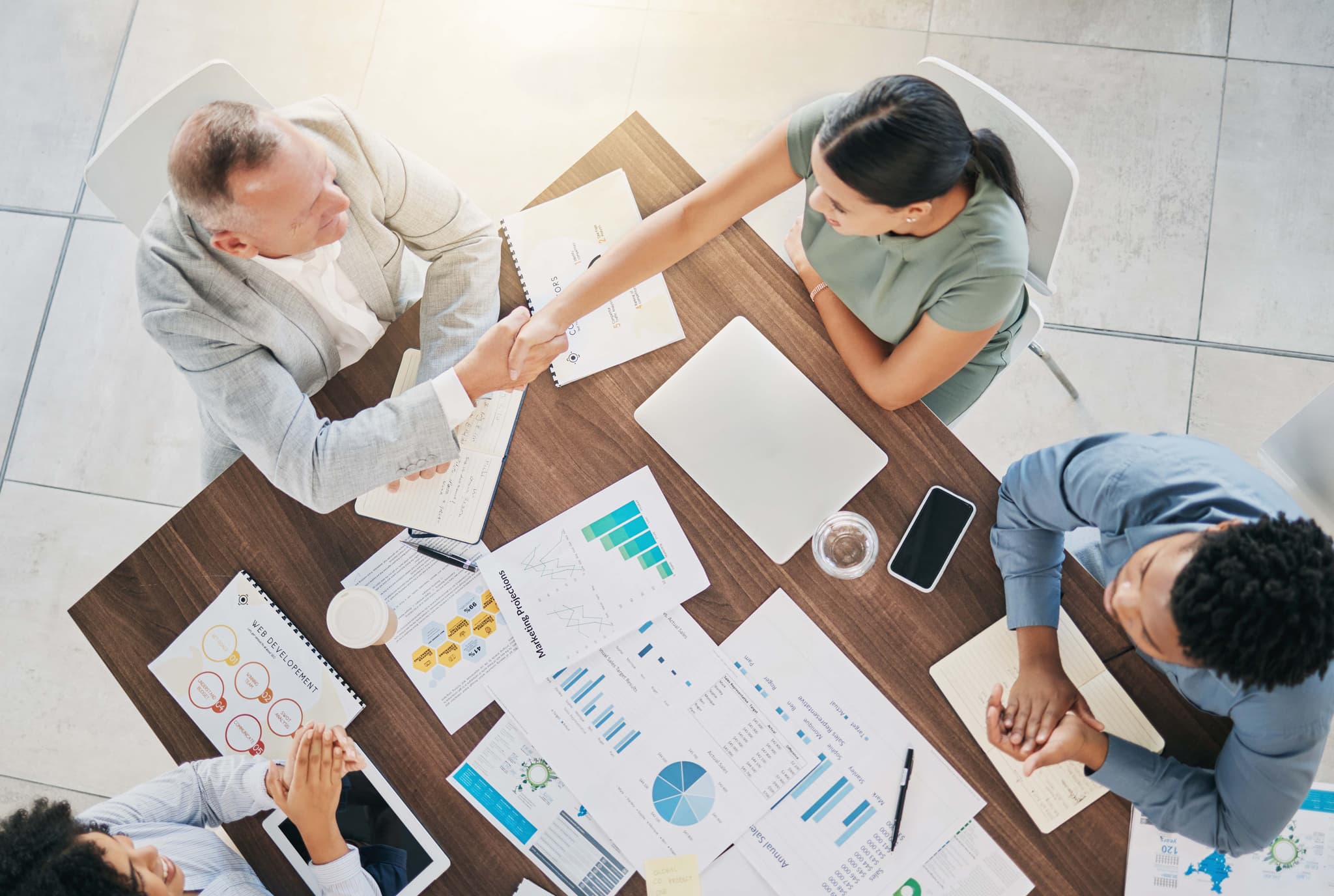 B2B Geschäftsabschluss – Handschlag über Konferenztisch mit Dokumenten, Charts und Laptops